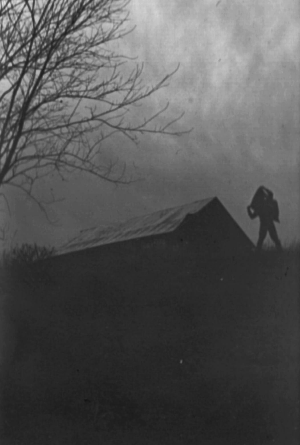 Grainy black and white photograph of a silhouetted figure with a flag on a hilltop beside a barn roof, under a bare tree and fog.