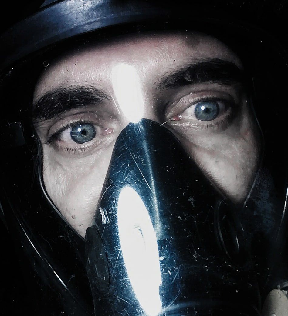 Close-up of a man's face inside a black respirator gas mask, his blue eyes lit by a bright glare across the visor.