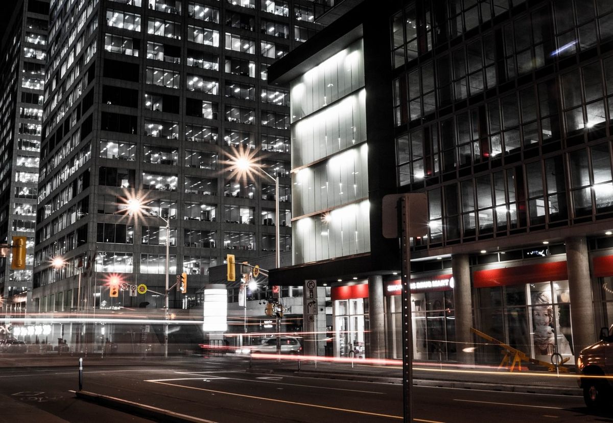 Night street scene of downtown office towers with lit windows, starburst street lamps, traffic lights and red light trails along the road.