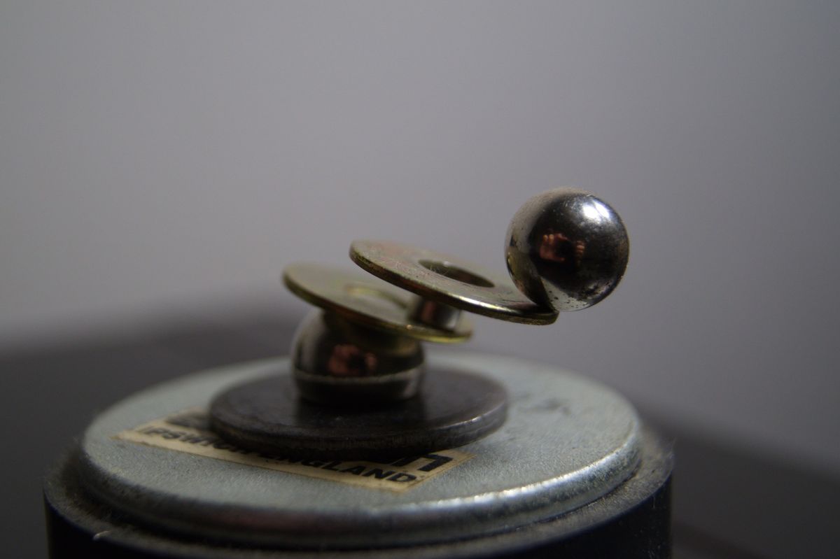 Close-up photograph of steel ball bearings and brass washers balanced on top of a metal cylinder against a soft grey background.