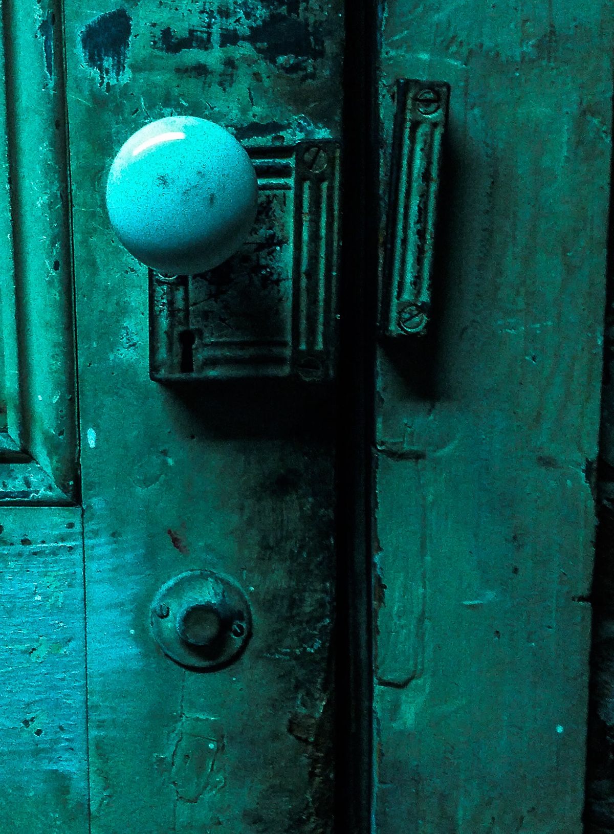 Close-up photograph of a weathered teal-painted door with a pale round doorknob, an old lock plate and scuffed, peeling paint.