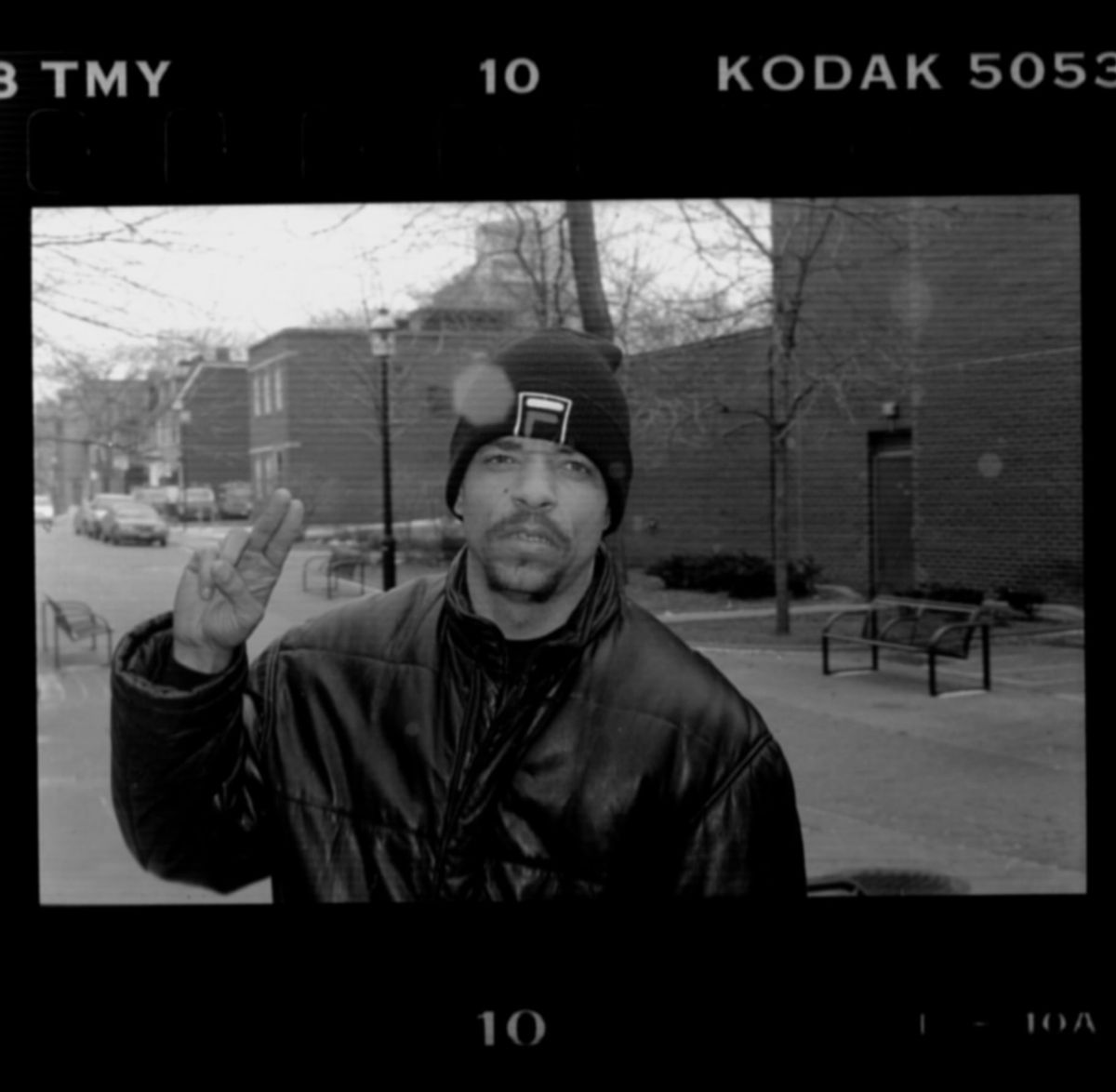 Black and white film photograph with Kodak frame edges of a man in a knit cap and leather jacket raising two fingers outdoors.
