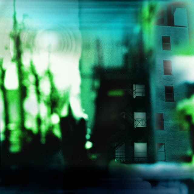 Blurred green and teal image of a dark brick apartment building with fire escapes beside glowing, smeared tree shapes.