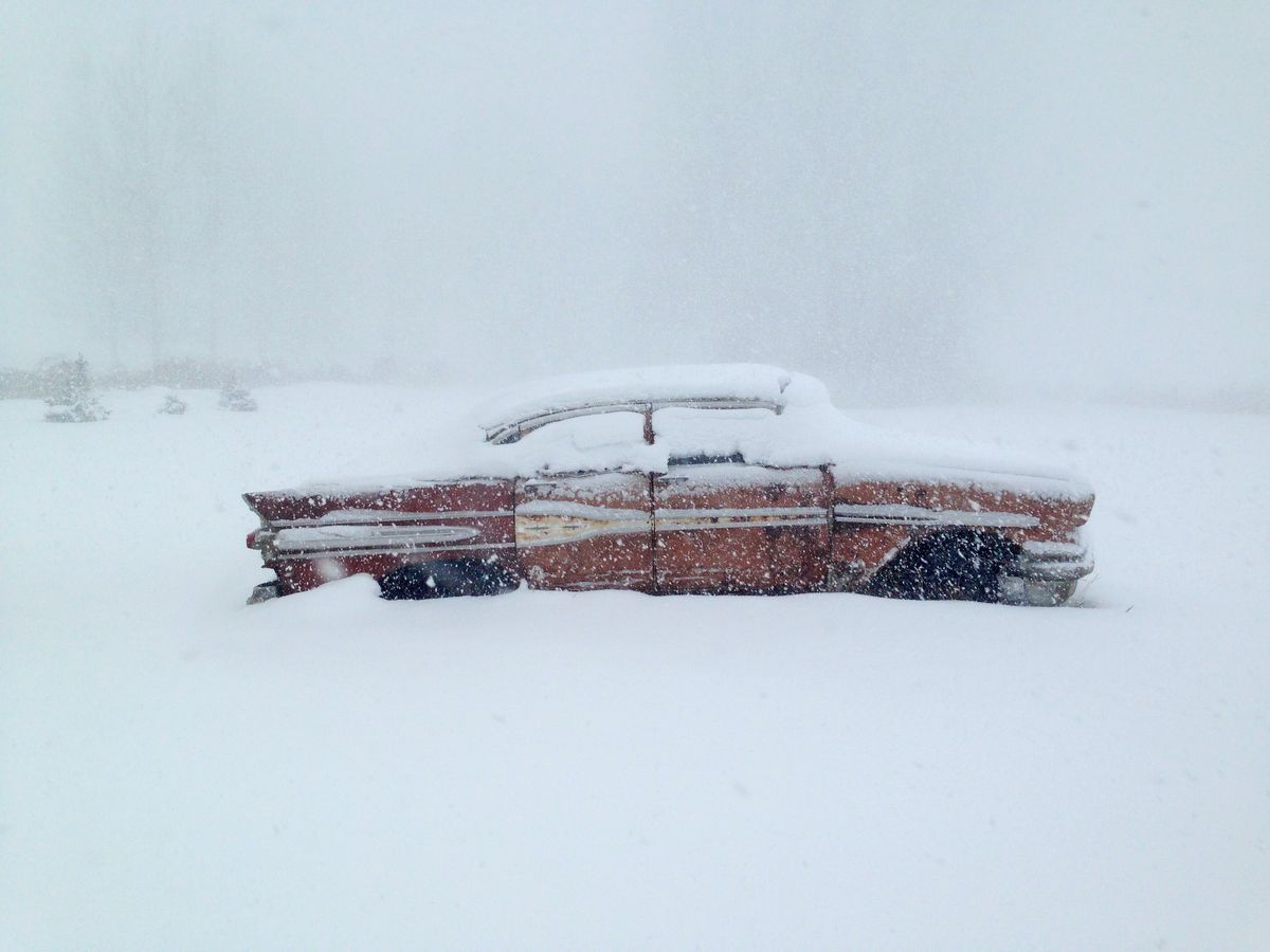 Photograph of a rusted red vintage car half buried in deep snow during a whiteout snowfall.