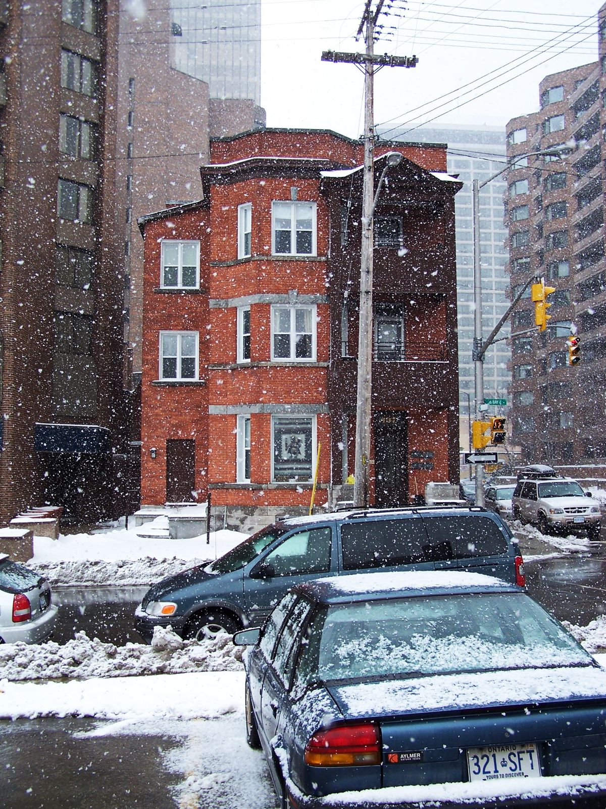 Snowy street photograph of a three-storey red brick house between high-rise towers, with parked cars, a utility pole and traffic lights.