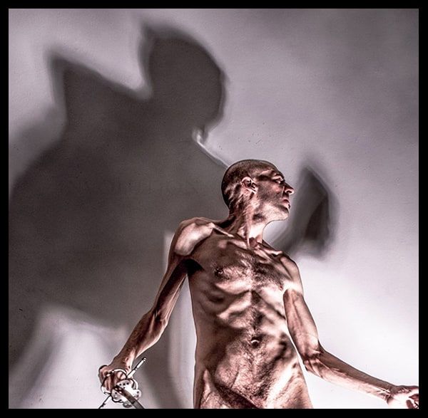 Photograph of a gaunt, shaven-headed nude man holding a sword hilt, lit warmly against a grey wall that carries his large shadow.