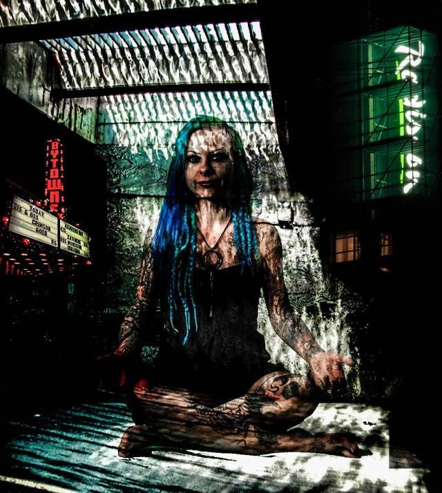 Tattooed woman with long teal braids sits cross-legged on a slatted floor beside a Bytowne theatre marquee and green neon lettering.