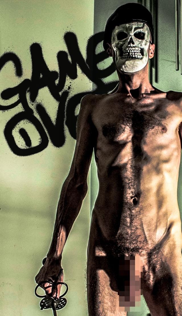 Nude man in a skull mask stands before a pale green wall sprayed with Game Over graffiti, holding a cord, his genitals pixelated.