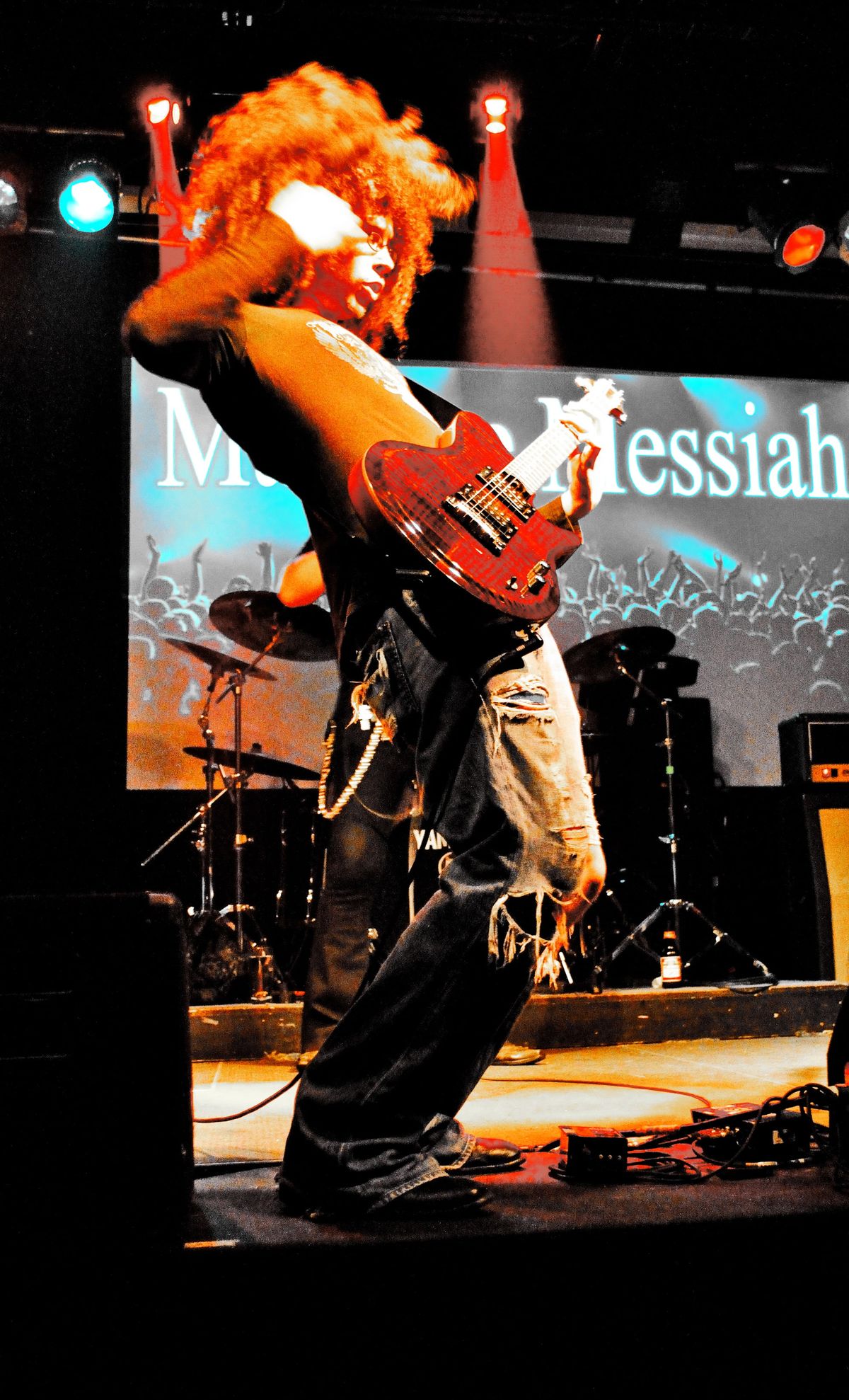 Concert photograph of a guitarist with a large red afro swinging a red guitar on a lit stage, a band name projected behind.