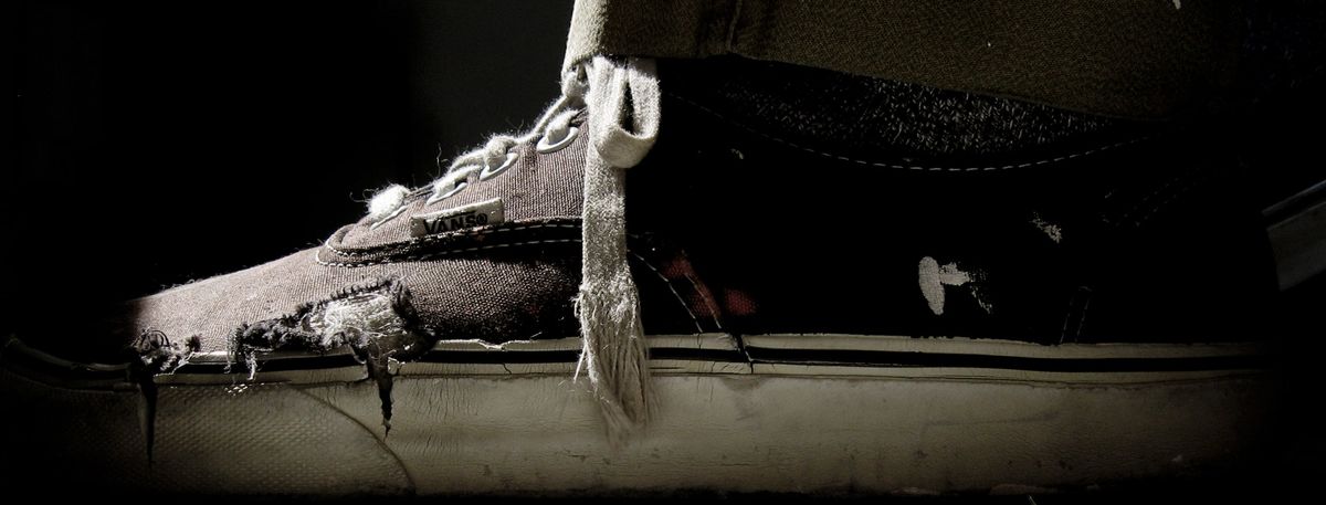 Close-up of a worn, frayed black canvas sneaker with a loose white lace, lit from the side against black.